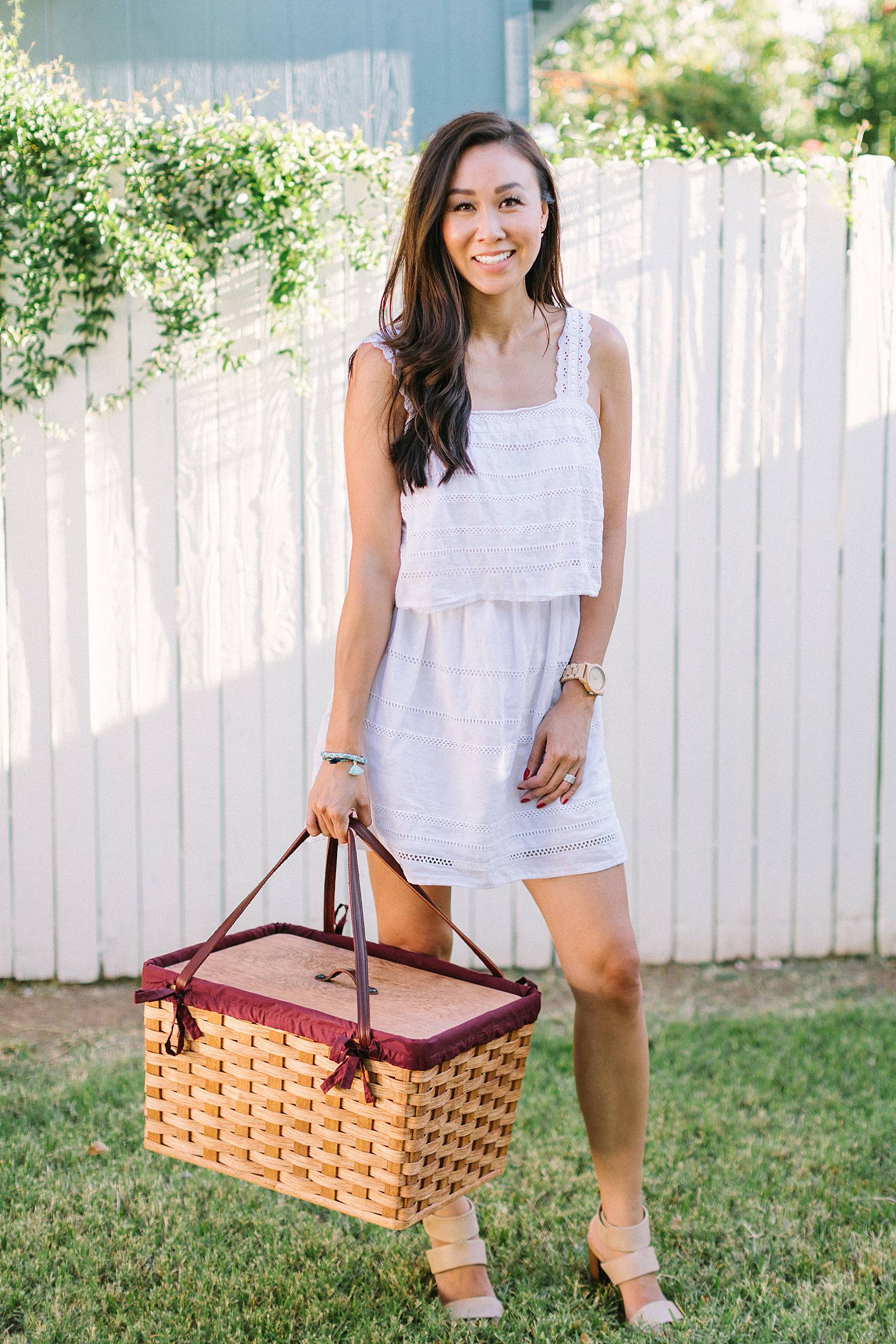 lifestyle blogger diana Elizabeth holding an amish made picnic basket in white dress infront of white fence standing on grass