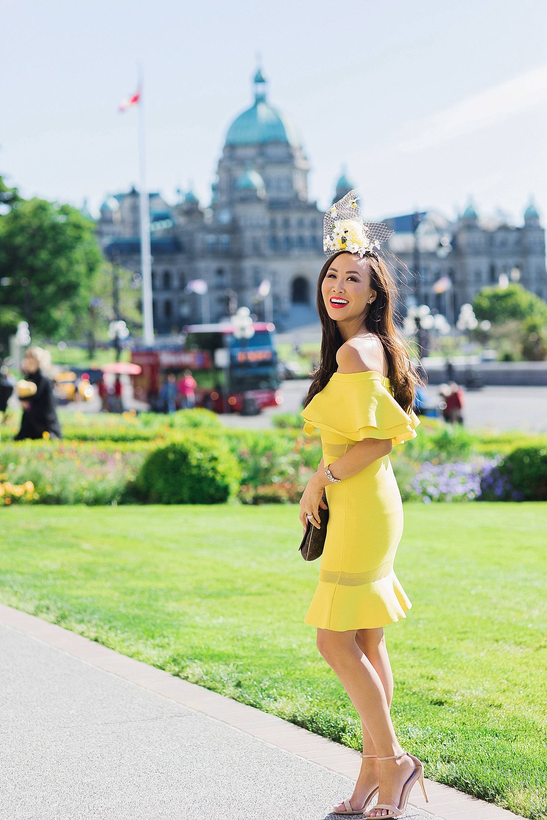 yellow fascinator hat bee hive high tea for bridal shower wedding celebration at empress hotel Victoria Canada wearing yellow off shoulder ruffle dress on blogger Diana Elizabeth standing in front of the Parliament building