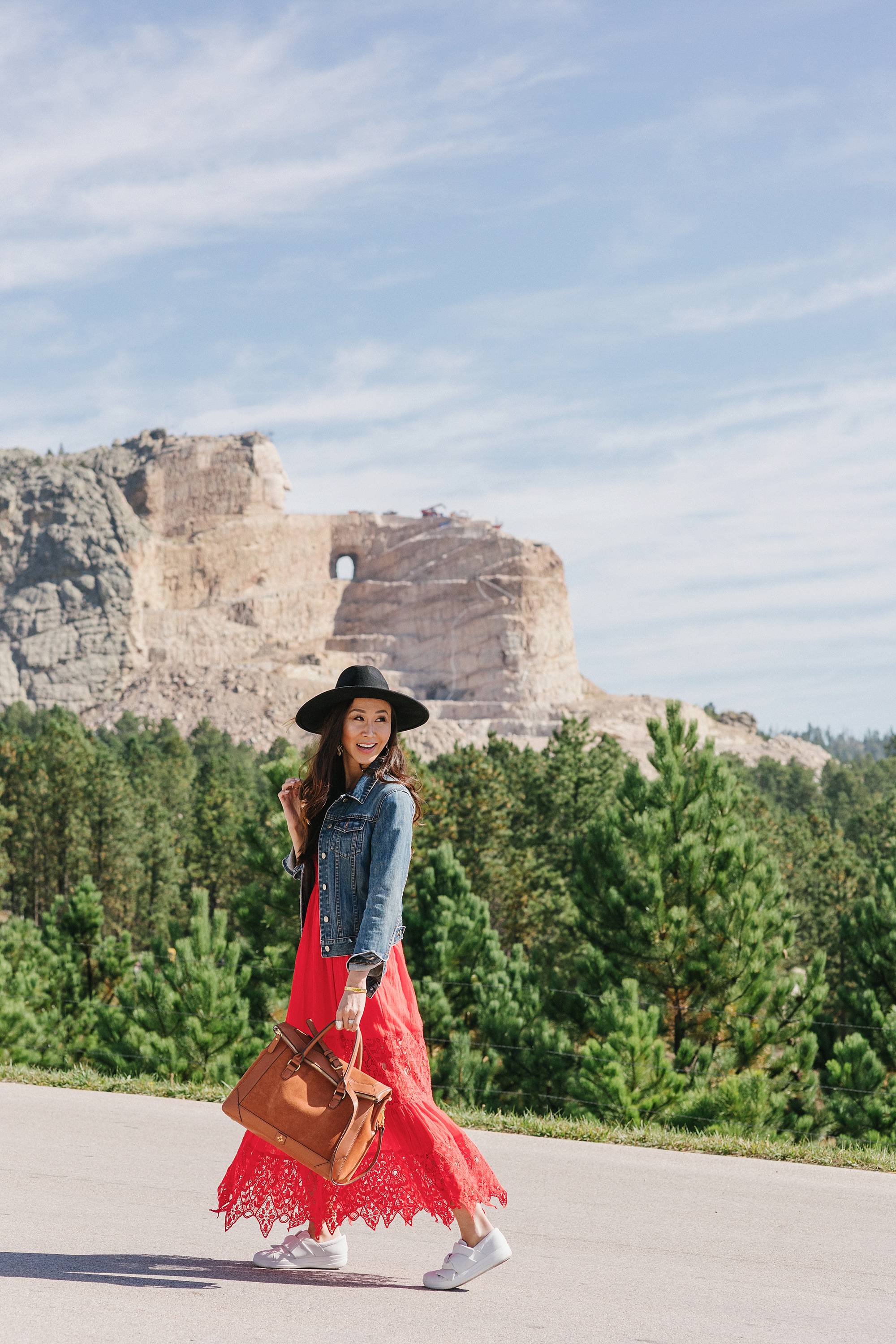 Crazy Horse Memorial in South Dakota - Diana Elizabeth Phoenix lifestyle blogger wearing red free people dress and wool hat and denim jacket posing and wearing white sneakers