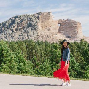 Crazy Horse Memorial in South Dakota - Diana Elizabeth Phoenix lifestyle blogger wearing red free people dress and wool hat and denim jacket posing and wearing white sneakers