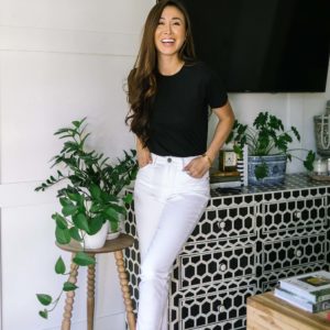 Diana Elizabeth phoenix lifestyle blogger in white pants blacks shirt leaning against black and white bone dresser by lots of house plants. jeans and top by Mott and bow
